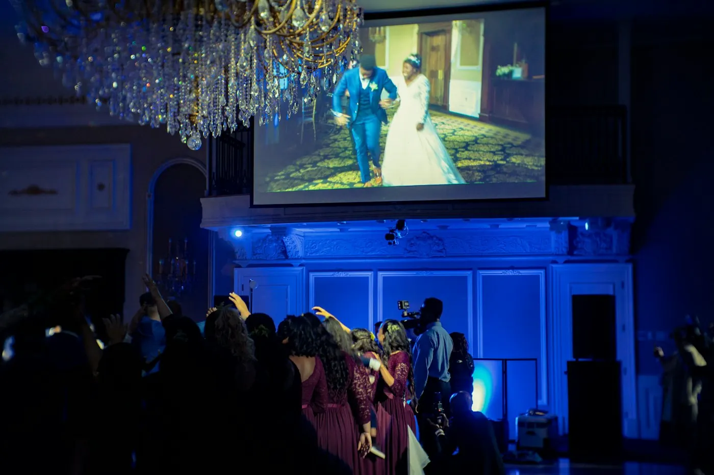 Une salle de réception de mariage : les invités devant un grand écran qui montre une photo des mariés